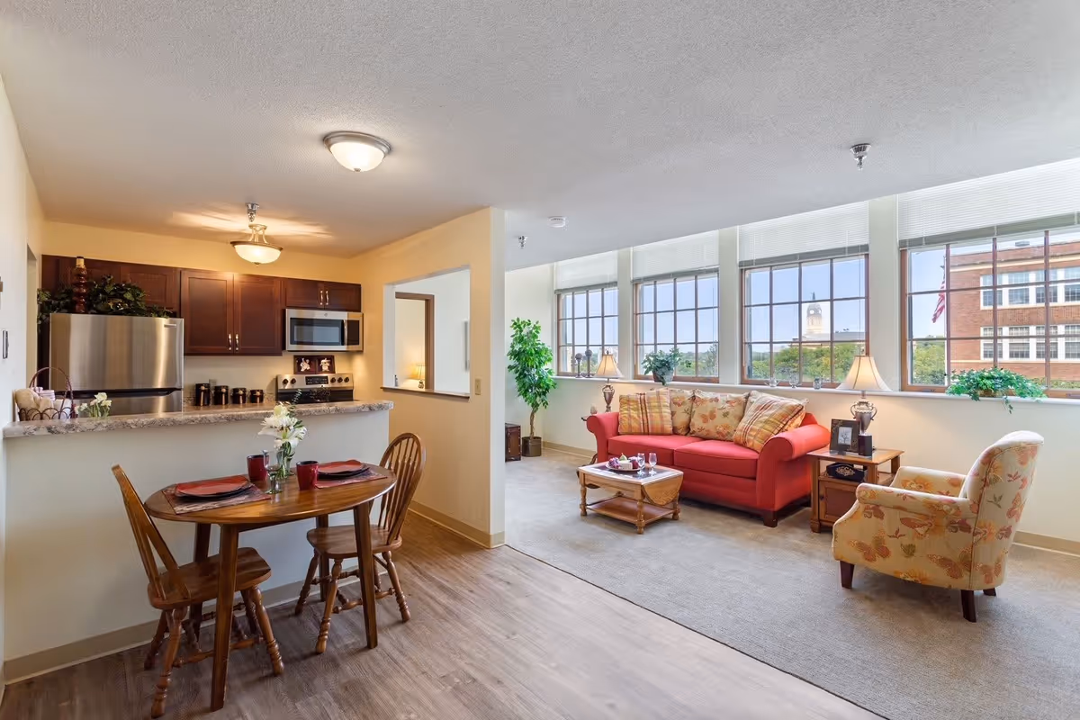 A bright and cozy living space in Old Main Village featuring a small wooden dining table with two chairs set with red placemats and cups. The kitchen area has dark wood cabinets, a stainless steel refrigerator, microwave, and stove. The living room has a red sofa with patterned cushions, a floral armchair, a wooden coffee table, and large windows letting in natural light with a view of a clock tower and a brick building outside.