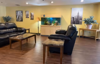 A cozy living room with two black leather sofas, a wooden coffee table, and a side table. There is a large fish tank on a wooden cabinet against a yellow wall, flanked by two potted plants on tables. The wall is decorated with framed black and white photographs and a floor lamp.