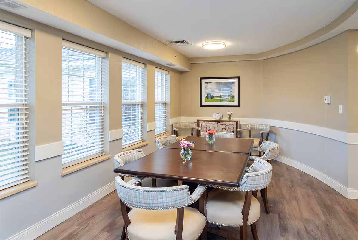 A bright dining room with a large wooden table surrounded by eight upholstered chairs. The room has multiple windows with white blinds, light beige walls, and wood flooring. There is a sideboard against the far wall with decorative items and a framed picture hanging above it. Small vases with flowers are placed on the table.