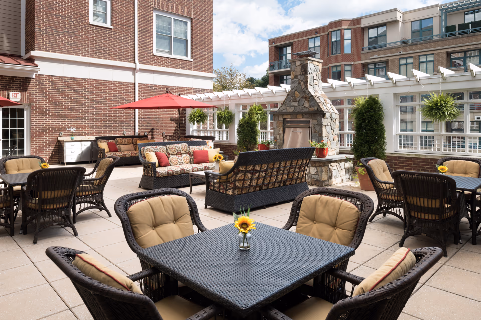 Outdoor patio area with multiple wicker tables and cushioned chairs, a stone fireplace, potted plants, and a red umbrella against a backdrop of brick buildings and a partly cloudy sky.