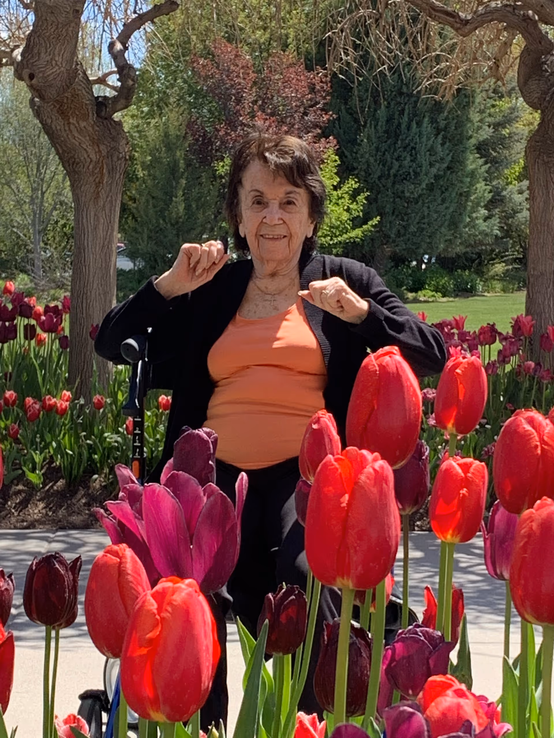 An elderly woman sitting in a wheelchair outdoors among vibrant red and purple tulips, with trees and greenery in the background. She is wearing an orange top and a black cardigan, smiling and raising her hands slightly.
