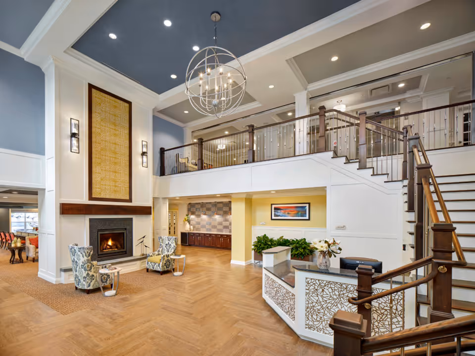 Spacious and elegant senior living facility lobby with a high ceiling, a modern chandelier, a fireplace with two patterned armchairs, a reception desk with decorative panels, a staircase with wooden handrails leading to an upper floor balcony, and a seating area visible in the background.