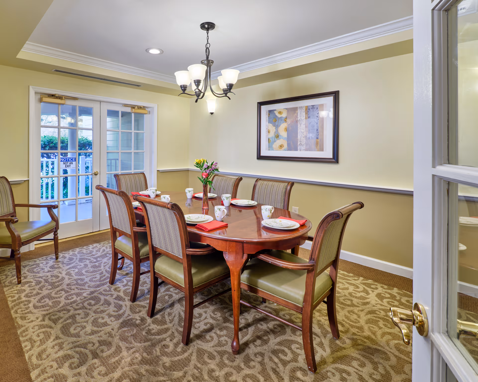 A dining room with a wooden table set for six, upholstered chairs, a chandelier, framed artwork, and French doors to the outside.