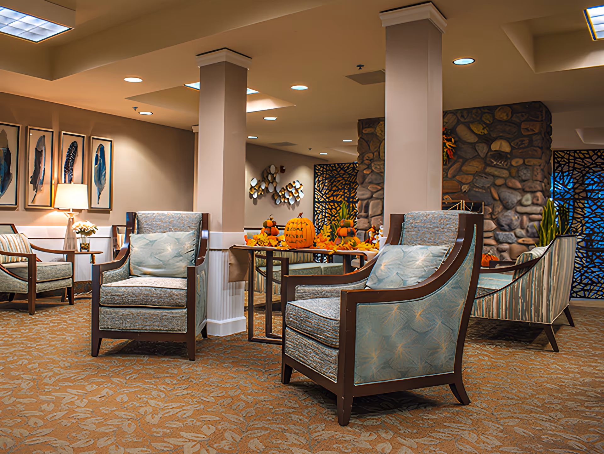 A cozy seating area in a senior living facility with several cushioned armchairs arranged around a small table decorated with autumn-themed pumpkins and leaves. The room features warm lighting, a stone accent wall, framed artwork of feathers, and a patterned carpet.