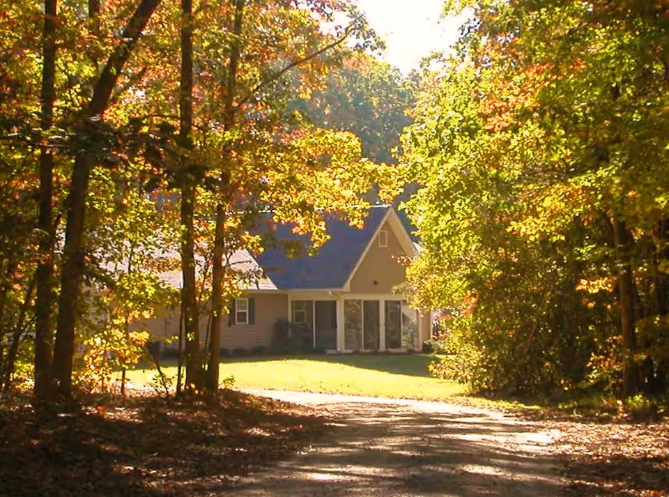 A house with a porch sits at the end of a tree-lined driveway surrounded by autumn foliage.