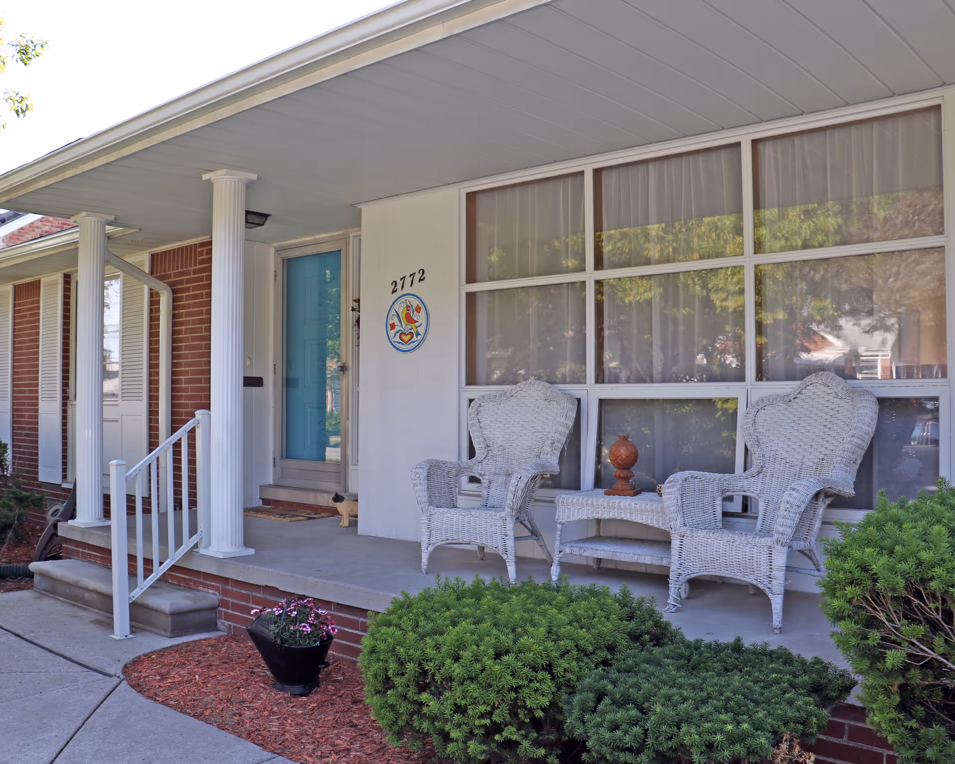 Front porch of a senior care facility with two white wicker chairs and a matching table. The porch has white columns and a blue front door with the number 2772 and a colorful circular sign on the wall. There are bushes and a flower pot near the porch.