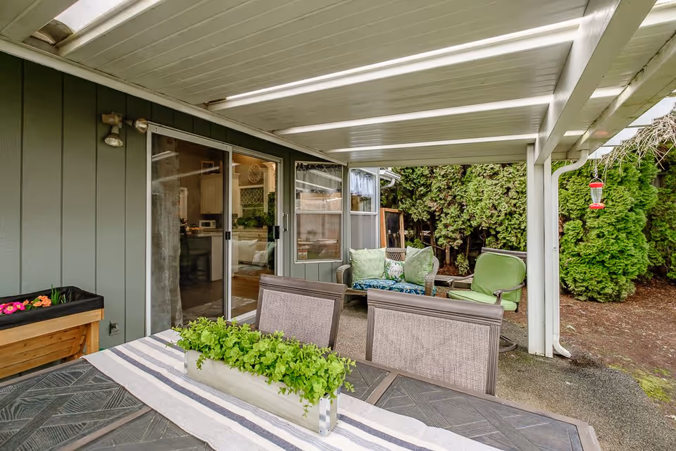Covered outdoor patio area with a table and chairs in the foreground, a planter with green plants on the table, and a seating area with green cushions and pillows near a sliding glass door leading inside. There are tall green bushes in the background and a hummingbird feeder hanging on the right side.