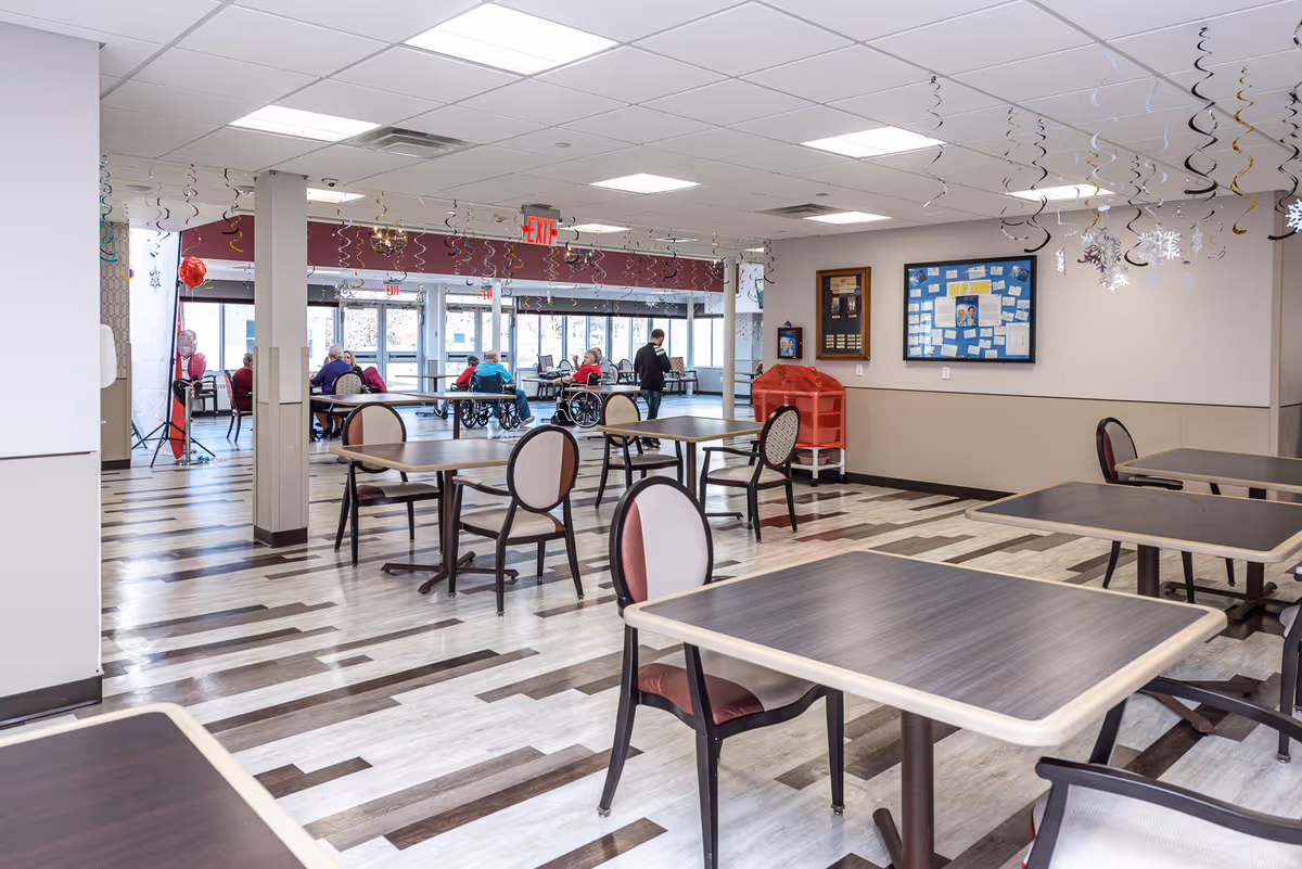 A spacious dining area in a senior living facility with multiple tables and chairs arranged neatly. Several elderly residents are seated near the large windows at the far end, some in wheelchairs. The ceiling is decorated with hanging spiral streamers and snowflake ornaments. The floor has a patterned wood design, and there are bulletin boards and a red storage bin against the wall.