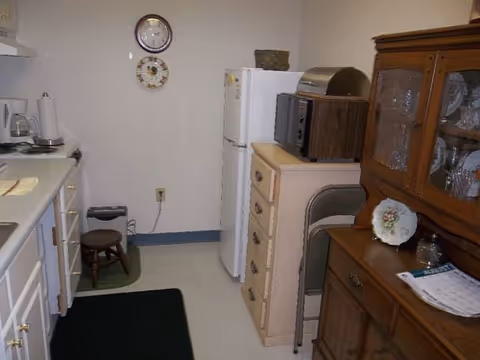 Small kitchen area with white cabinets and countertop on the left, a white refrigerator at the back, a microwave and toaster oven on top of a small chest of drawers, a wooden hutch with glassware on the right, and a small stool on a green mat near the wall. A clock and decorative plate hang on the wall above the refrigerator.