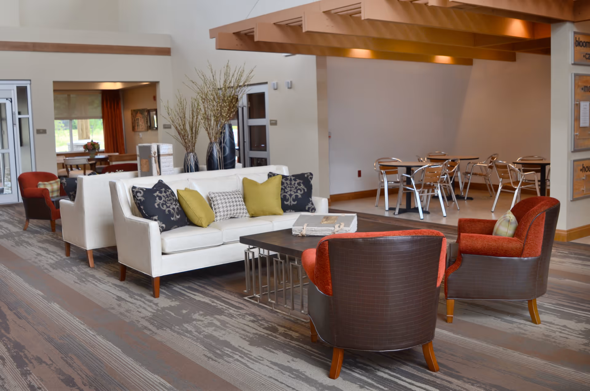 A spacious and well-lit common area in a senior living facility featuring a white sofa with decorative pillows, two red armchairs, a dark wooden coffee table, and a dining area with metal chairs and tables in the background. The room has a modern design with a patterned carpet and large windows letting in natural light.