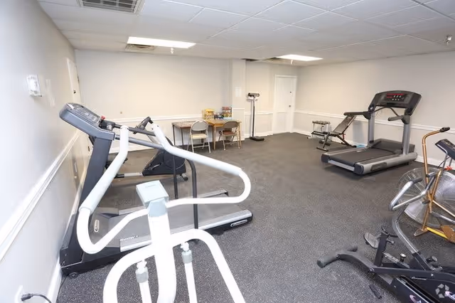 A small fitness room with exercise equipment including treadmills, an elliptical machine, and stationary bikes. There is a table with two chairs against the far wall, and the room has white walls and a gray carpeted floor.