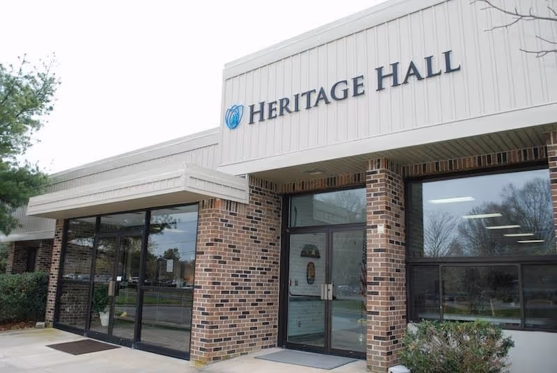 Exterior front entrance of Heritage Hall Nassawadox building with brick walls, large glass windows, and double glass doors under a beige overhang.