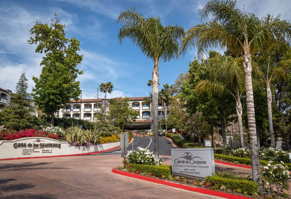 Entrance driveway to Casa de las Campanas, a continuing care retirement community, with palm trees, landscaped flower beds, and a multi-story building in the background under a blue sky.