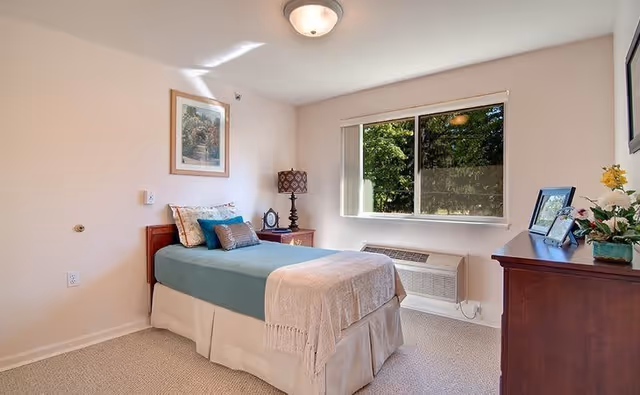 A neatly arranged single bedroom in a senior living facility with a bed covered in teal and beige bedding, a wooden nightstand with a lamp and clock, a window showing greenery outside, and a wooden dresser with framed photos and a flower arrangement.