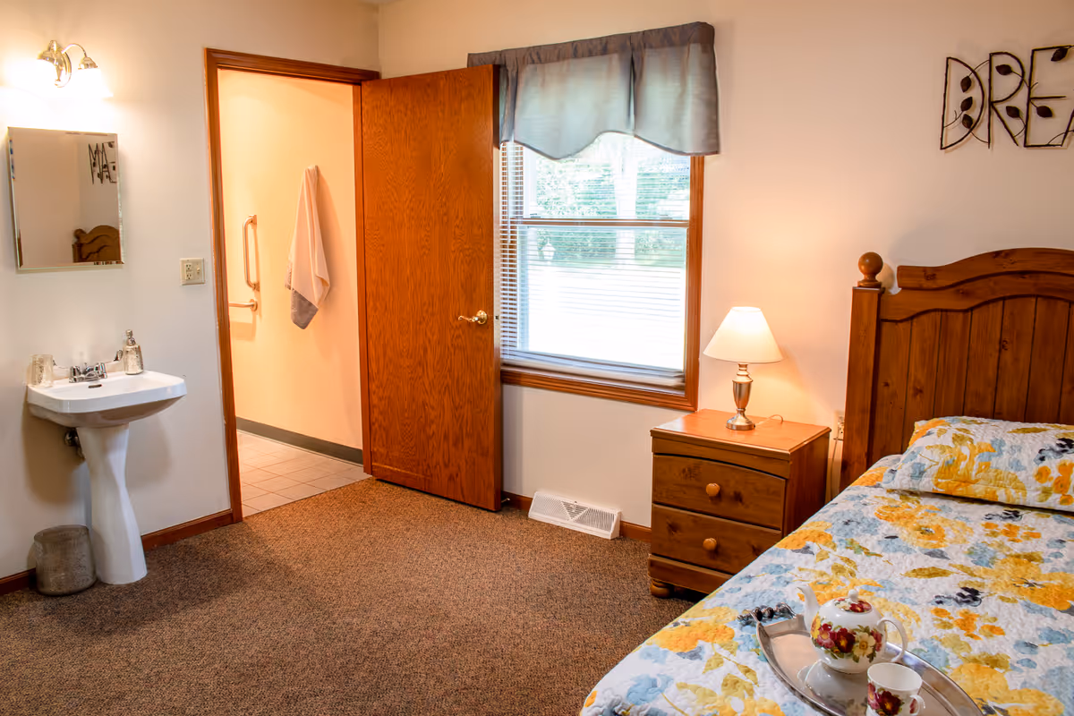 A cozy bedroom in a senior living facility featuring a wooden bed with a floral quilt, a wooden nightstand with a lamp, a window with a blue valance, and an open door leading to a bathroom area with a towel hanging on the wall. A white pedestal sink with a mirror and light fixture is visible on the left side of the room.