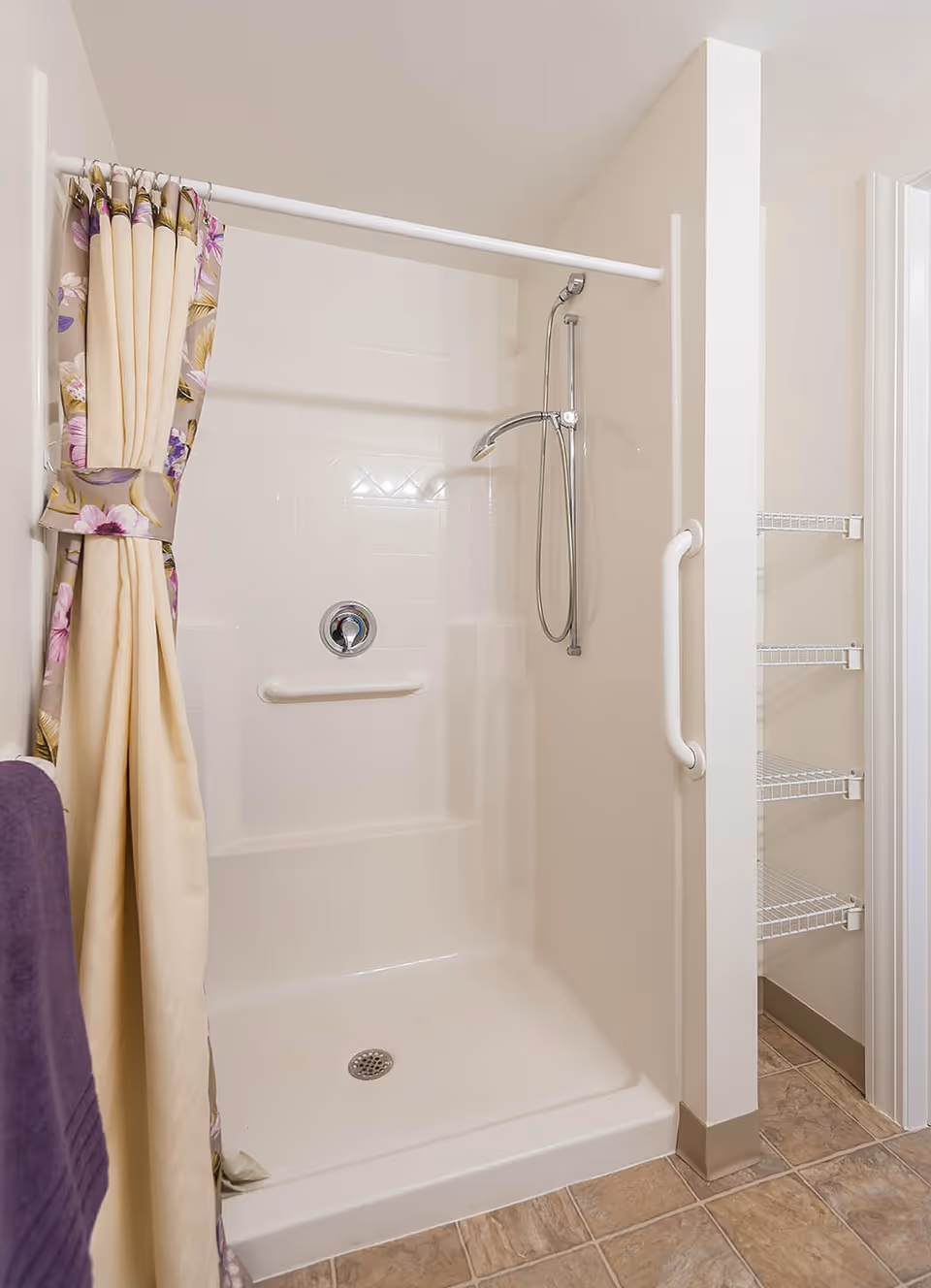 A clean, accessible shower area with a white shower base and walls, a handheld showerhead mounted on a vertical bar, a grab bar on the wall, and a floral-patterned shower curtain tied back. To the right, there are wire shelves for storage and a white grab bar on the wall. The floor is tiled in a beige color.