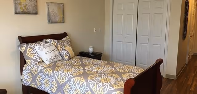 A bedroom with a wooden bed frame featuring a patterned bedspread in gray, white, and yellow. There are three pillows on the bed, one with the word 'SNOOZE' printed on it. Next to the bed is a small black nightstand with a white lamp. The room has light-colored walls, two framed artworks above the bed, a white double-door closet, and a wooden floor.