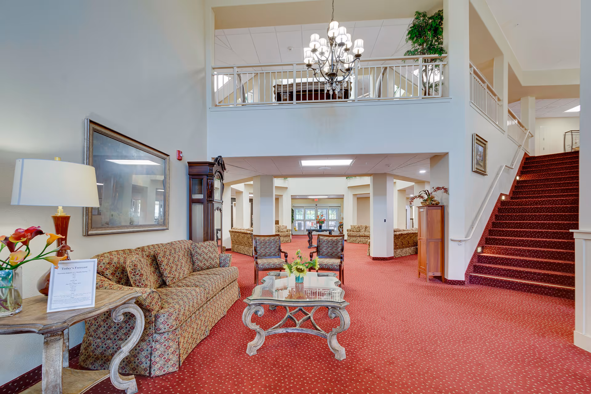 Spacious two-story senior living lobby with patterned sofas, a glass coffee table, chandelier, and red carpeted stairs.