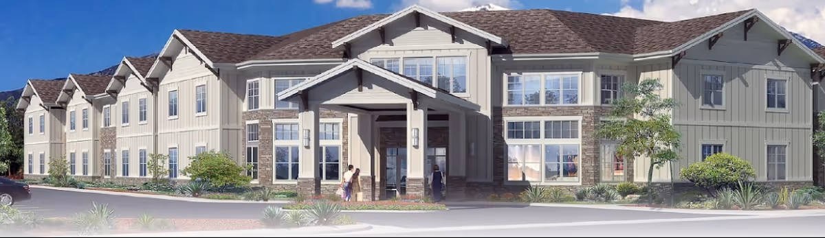 Exterior front view of a two-story senior living facility building with beige siding, stone accents, multiple windows, and a covered entrance. There are a few people near the entrance and a car parked on the left side. The sky is blue with some clouds.