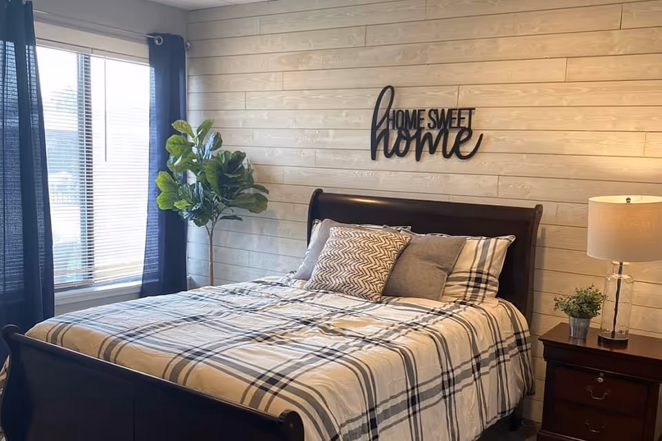 A cozy bedroom with a large bed featuring plaid bedding and multiple pillows. Above the bed is a decorative sign that reads 'Home Sweet Home'. To the right of the bed is a wooden nightstand with a lamp and a small potted plant. To the left, there is a large window with dark curtains and a tall green plant in the corner. The walls are covered with light-colored wooden panels.