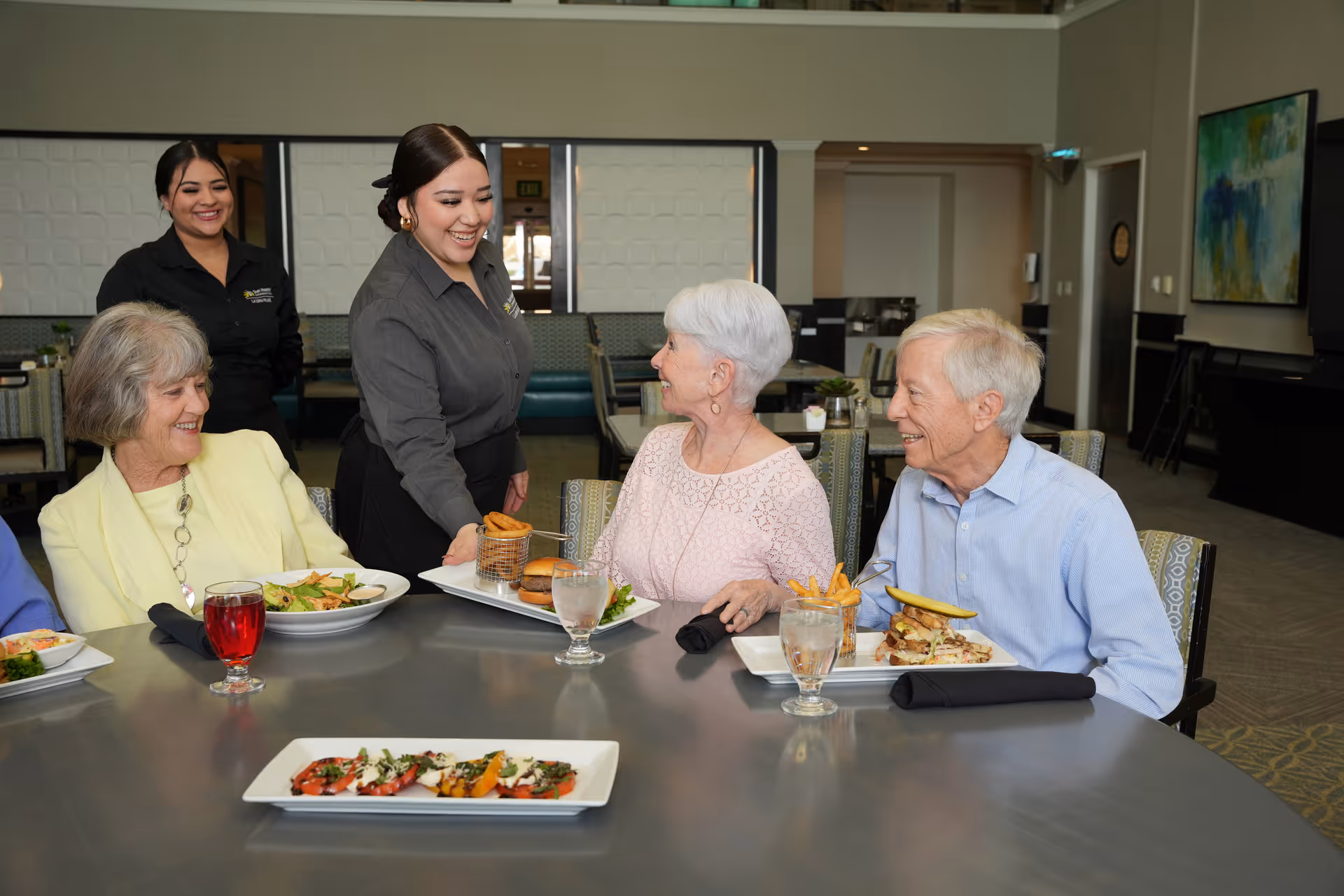 Two elderly women and one elderly man sitting at a dining table with plates of food and drinks, smiling and interacting with two female staff members who are serving food in a well-lit dining room.