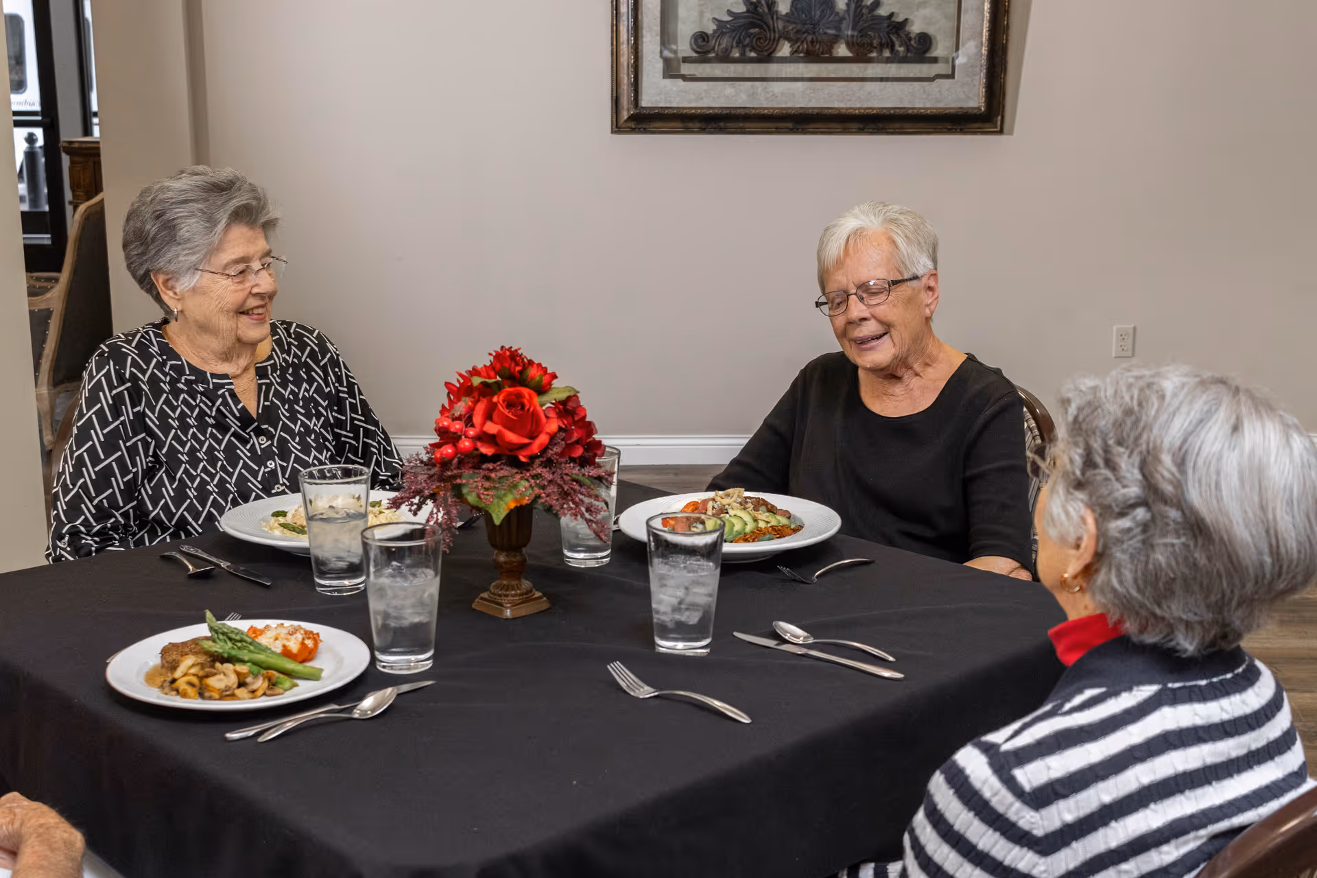 Three elderly women sit around a dining table with plates of food and a floral centerpiece, smiling and conversing.