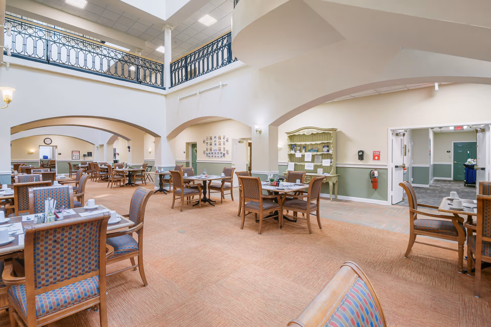A spacious dining room in a senior living community with multiple wooden tables and chairs arranged neatly. The room features high ceilings with a second-floor balcony railing, arched doorways, and soft lighting. Tables are set with cups, plates, and utensils, ready for a meal service.