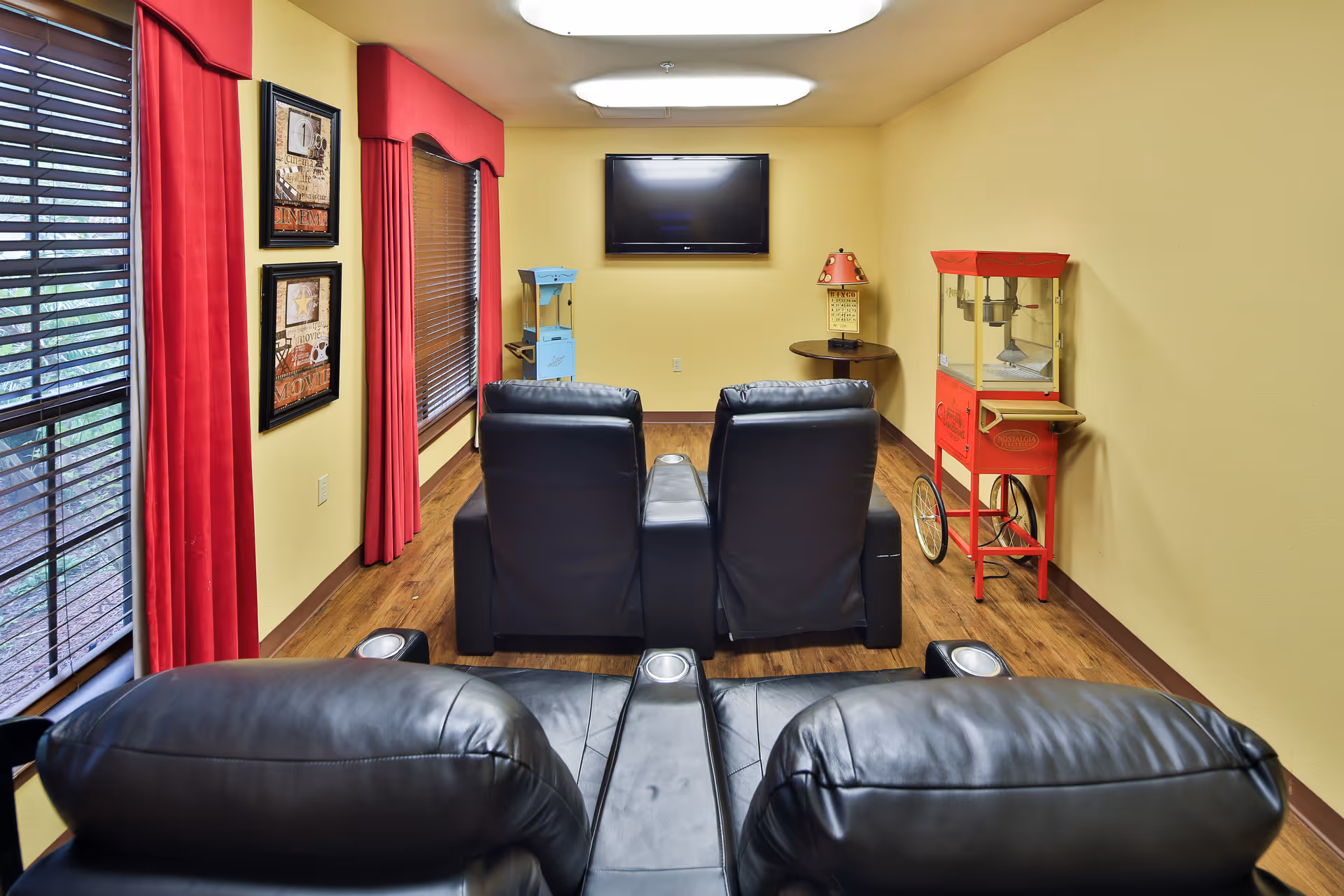 A small entertainment room with four black leather recliner chairs facing a wall-mounted flat screen TV. The room has yellow walls, wooden flooring, and two windows with red curtains on the left side. There is a red vintage-style popcorn machine on the right side and a small round table with a lamp next to it. Two framed pictures hang on the wall between the windows.