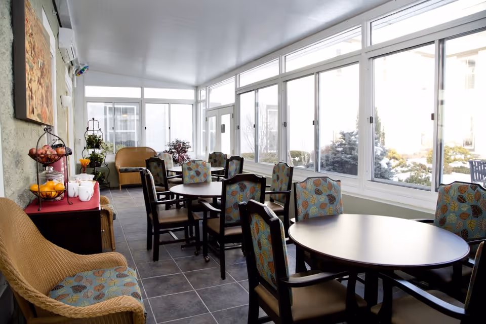 Bright enclosed sunroom dining area with round tables and patterned chairs along large windows.