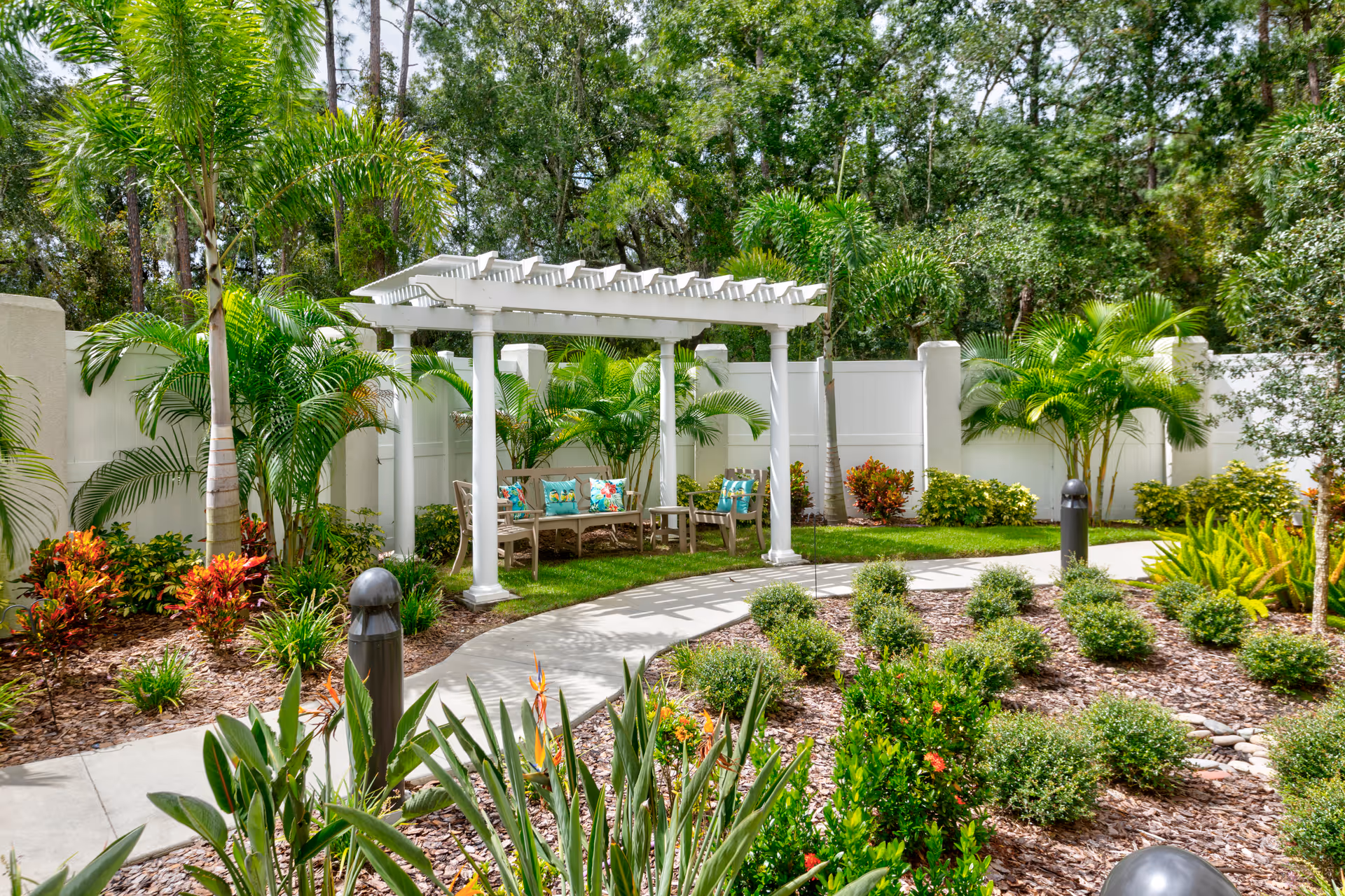 A peaceful outdoor garden area with a white pergola covering a seating area that includes benches with colorful cushions. The garden is surrounded by lush green plants, palm trees, and a white fence, with a curved concrete pathway leading through the landscaped space.