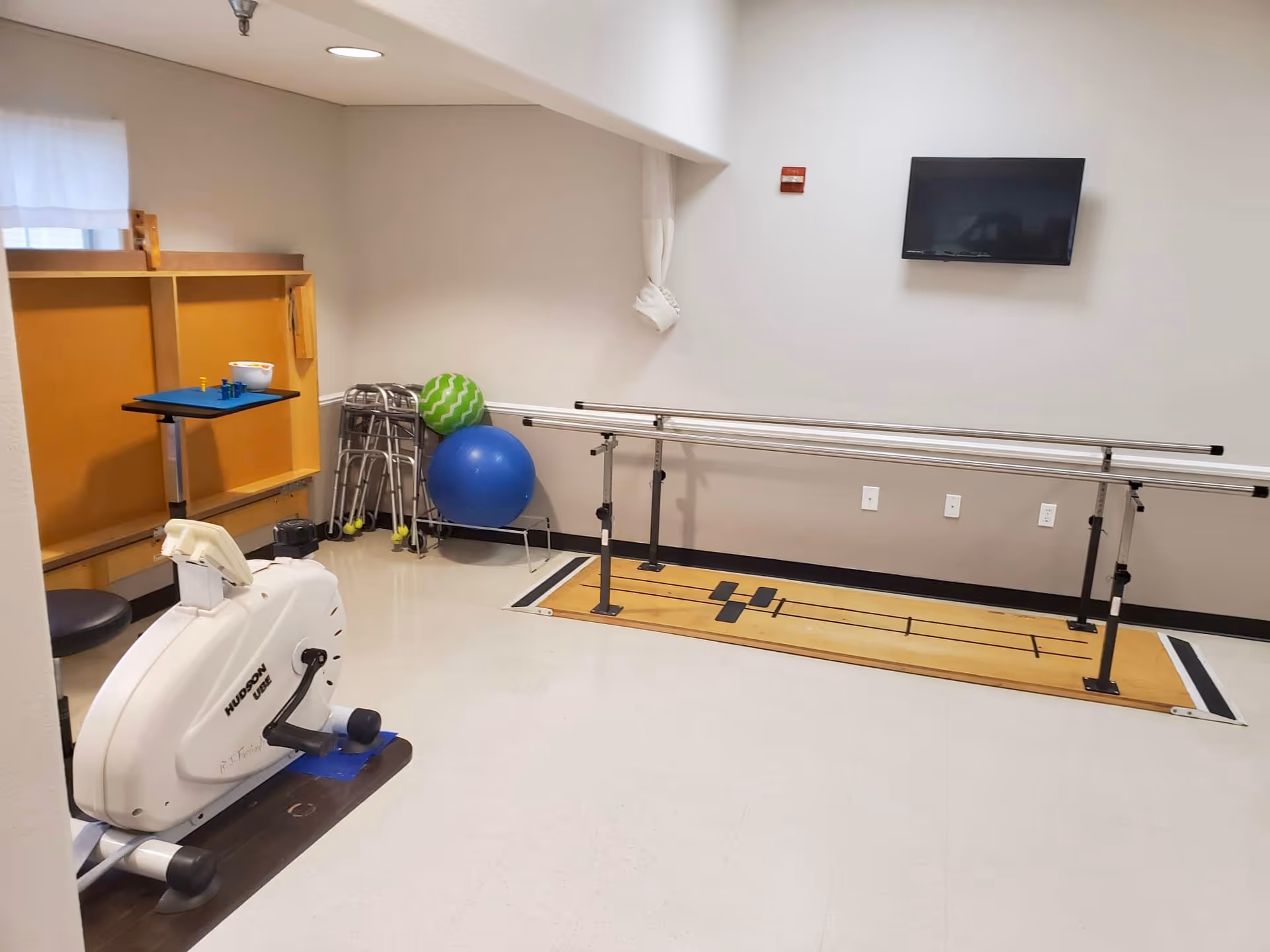A rehabilitation room with parallel bars for walking exercises, a stationary exercise bike, several walkers, and two exercise balls. A flat-screen TV is mounted on the wall, and the room has a clean, bright appearance with light-colored walls and flooring.