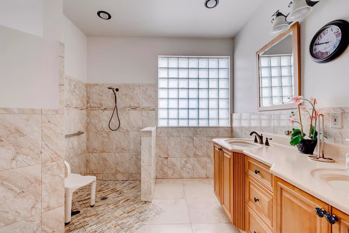 Spacious tiled bathroom with a curbless walk-in shower and white shower chair, a glass-block window, and a double-sink wooden vanity with mirror.