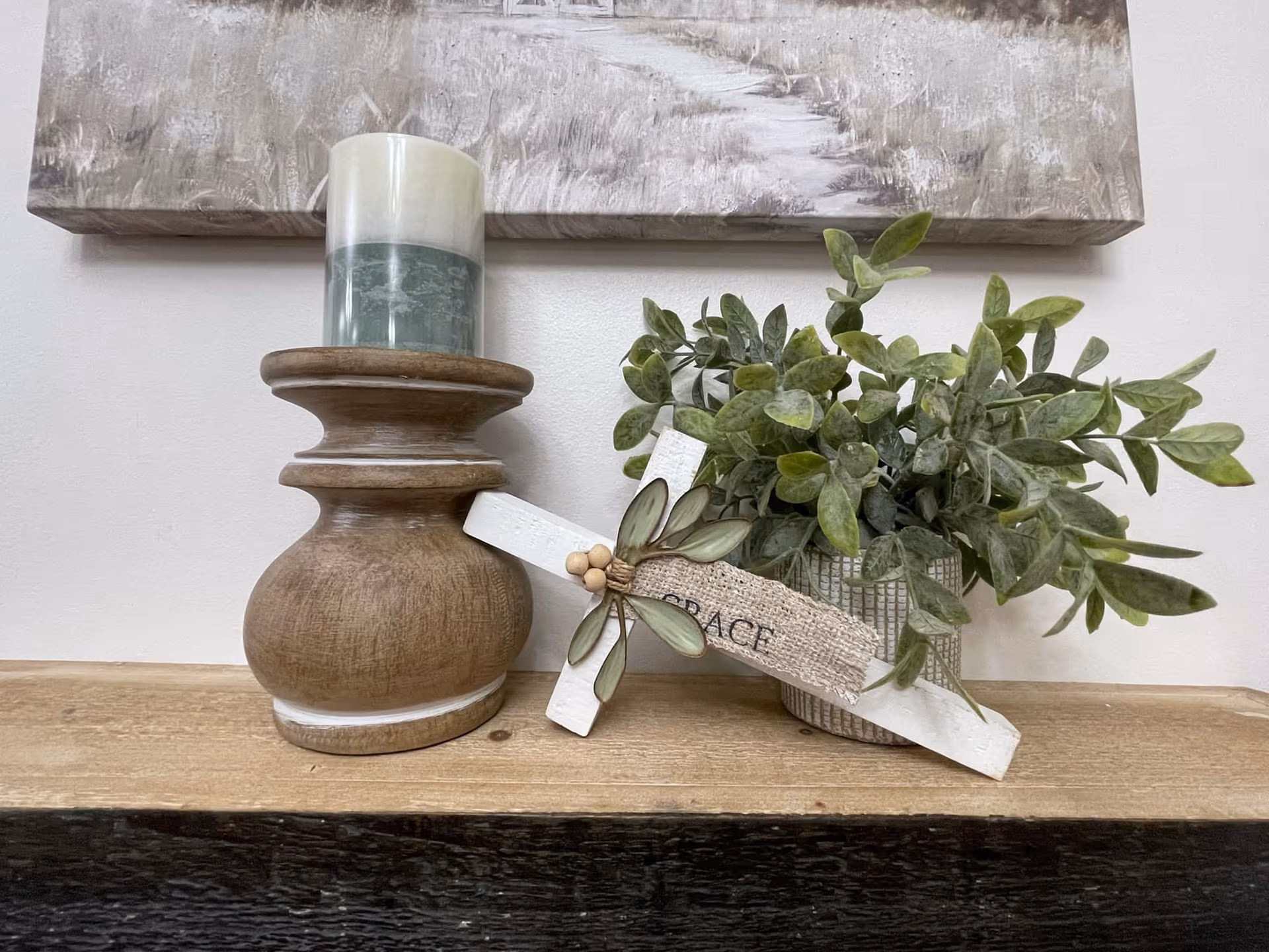 A decorative arrangement on a wooden surface featuring a two-toned candle on a wooden holder, a small potted plant with green leaves, and a white wooden cross with a burlap ribbon that says 'GRACE'. A painting with a muted color palette is partially visible on the wall behind the items.