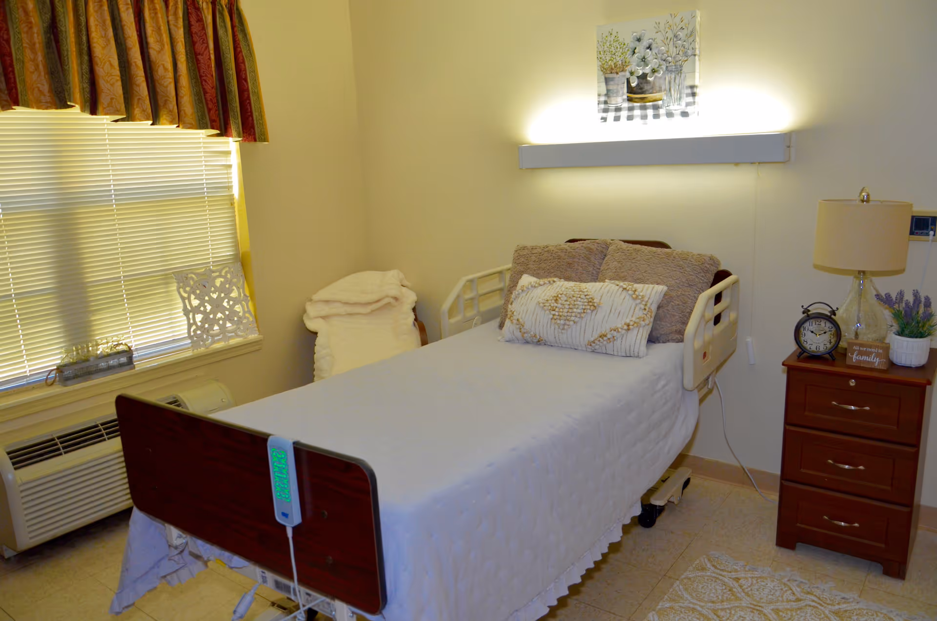 A tidy nursing facility bedroom with a single hospital bed, bedside table with lamp and clock, window with blinds, and wall light above the bed.