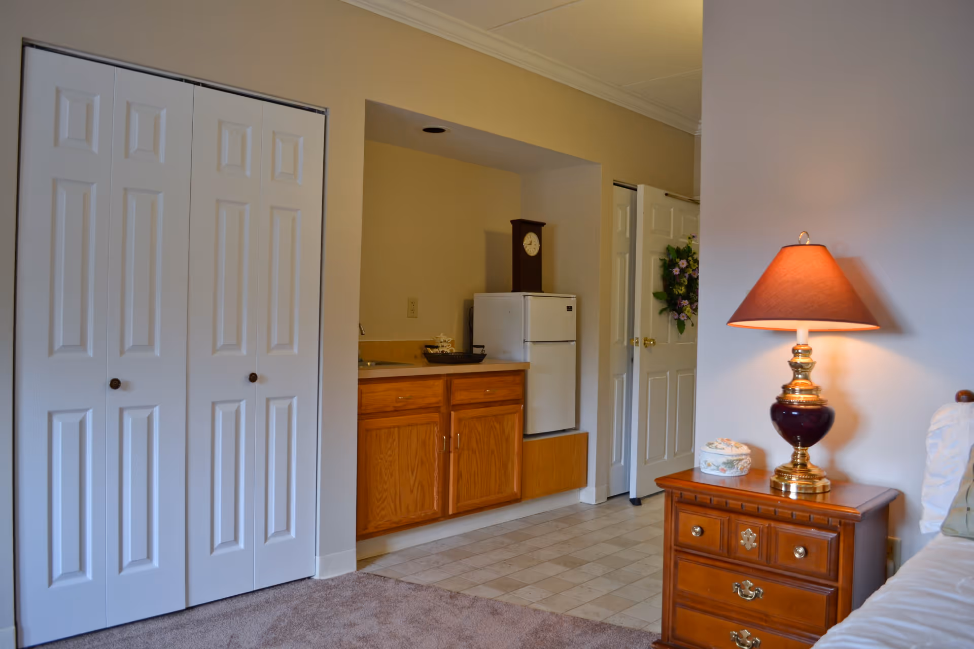 Interior studio-style room with a small kitchenette, white closet doors, a wooden nightstand with a lamp, and a partial view of a bed.