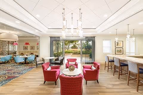 A bright and spacious common area in a senior living facility featuring a seating arrangement with four red armchairs around a round white table on a wooden floor. To the right, there is a long counter with bar stools. Large sliding glass doors open to an outdoor garden area with trees and greenery. The ceiling has modern light fixtures, and the room is decorated with framed artwork and patterned curtains.