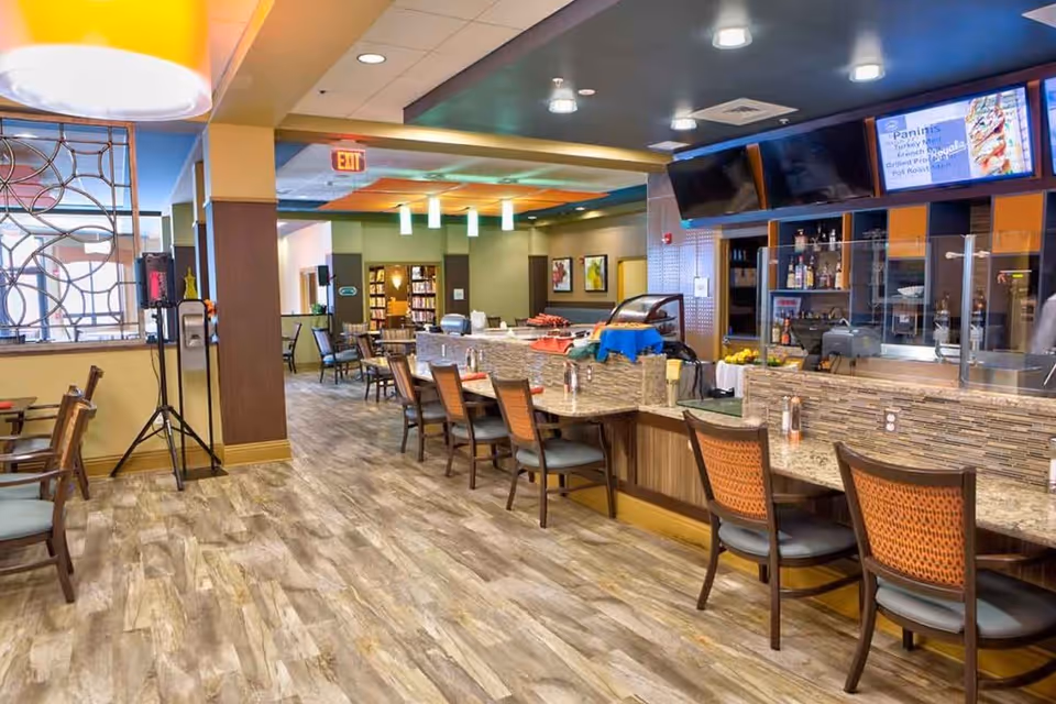 Interior view of a senior living community dining area with a long counter and several chairs. The counter has a stone facade and is equipped with condiments and a food display case. There are multiple TV screens mounted above the counter displaying menu items. The room has wood-look flooring, modern lighting fixtures, and a warm color palette. In the background, there is a seating area with tables and chairs and a bookshelf.