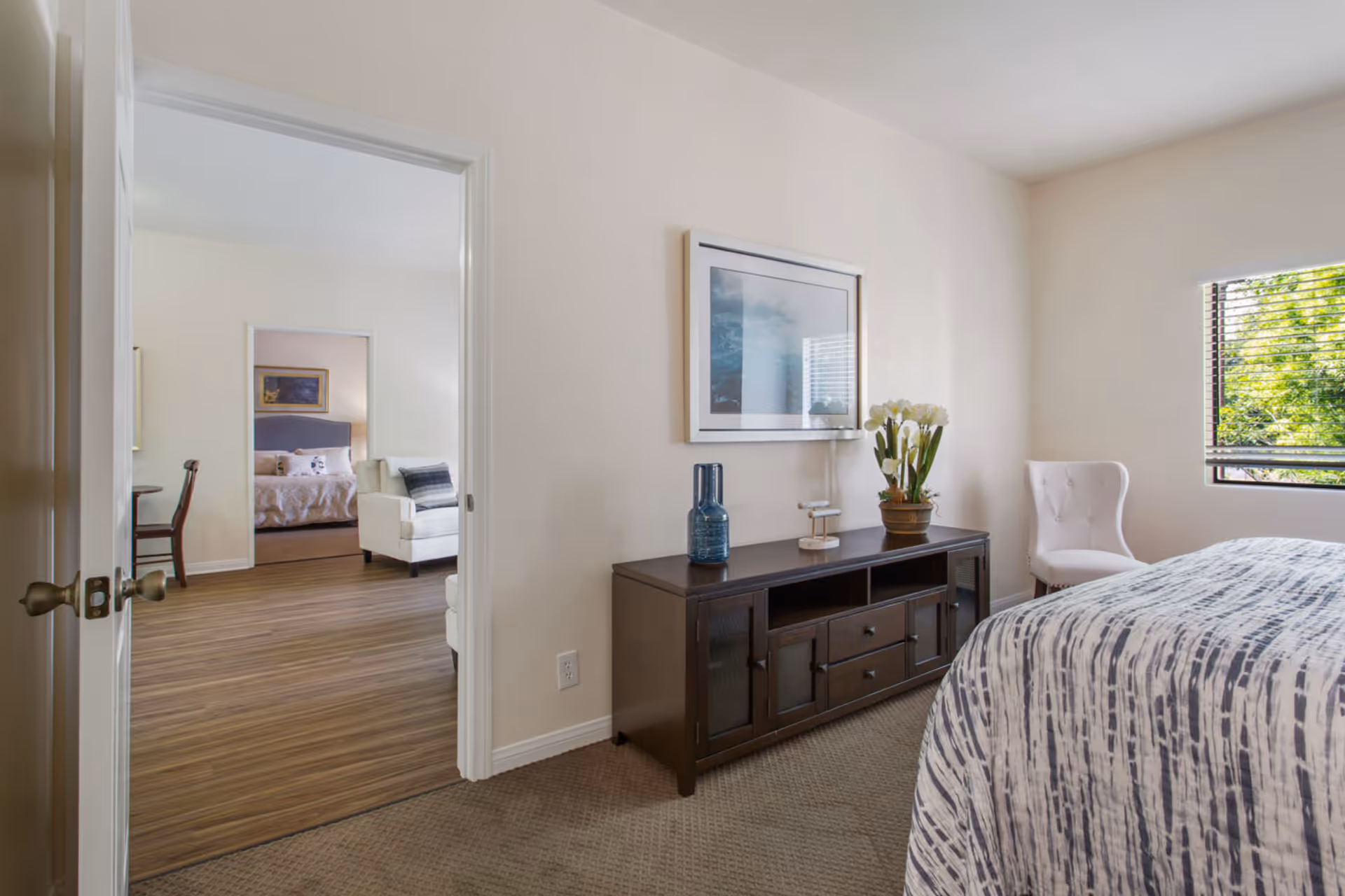 Interior view of a bedroom with a bed covered in a patterned bedspread, a white upholstered chair near a window with blinds, and a dark wooden dresser with decorative items including a blue vase and a potted plant. The room has beige walls and carpeted floor. Through an open door, a living area with wooden flooring, a white armchair, and a glimpse of another bedroom with a bed and framed artwork is visible.