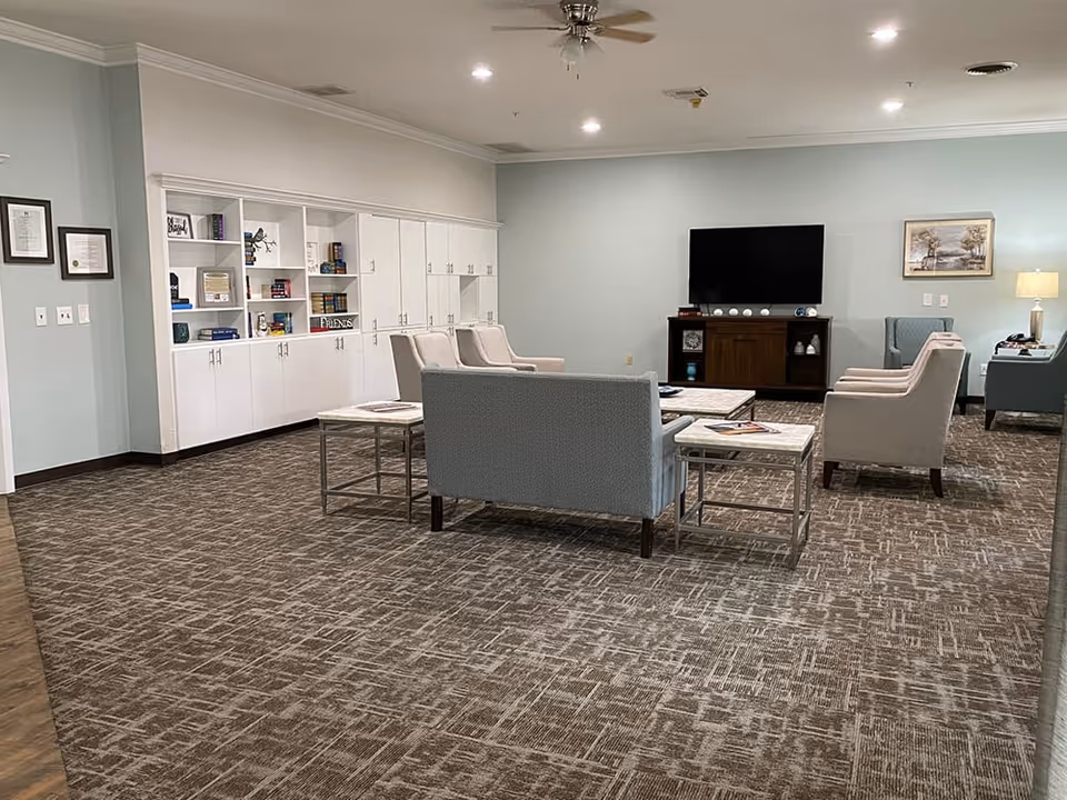 A senior living facility common area with a carpeted floor, light blue walls, and recessed ceiling lights. The room features a seating arrangement with a blue loveseat, beige armchairs, and side tables. A large flat-screen TV is mounted on a wooden stand against the far wall, with decorative items and a framed picture hanging nearby. Built-in white cabinets and shelves filled with books and decor line one wall.