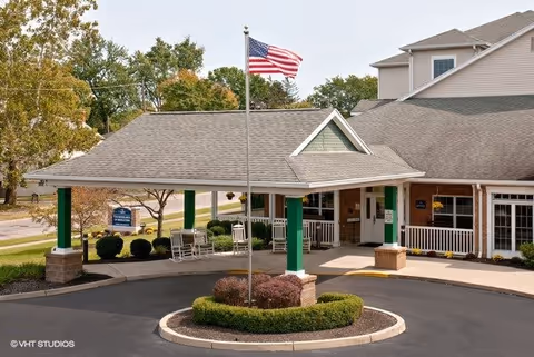 Entrance of a senior living facility with a covered driveway supported by green pillars, an American flag on a flagpole in the center of a circular landscaped area, and a building with beige siding and white trim in the background.