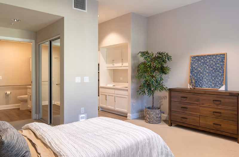 Bedroom in a senior living facility with a dresser, plant, and open doorway to a bathroom.