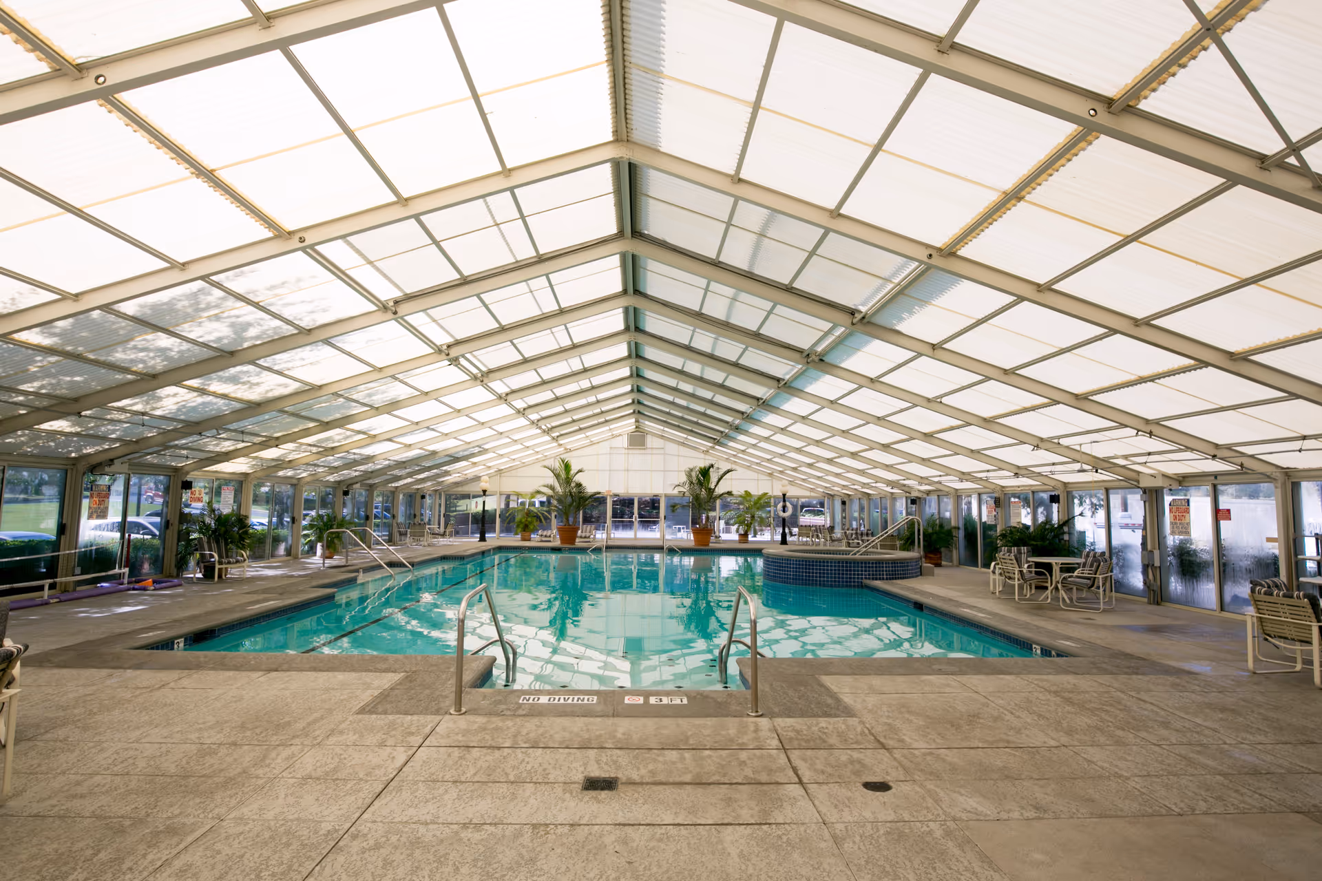 Indoor swimming pool area with a clear roof and glass walls, surrounded by potted plants and seating areas with tables and chairs. The pool has metal handrails and a hot tub adjacent to it.
