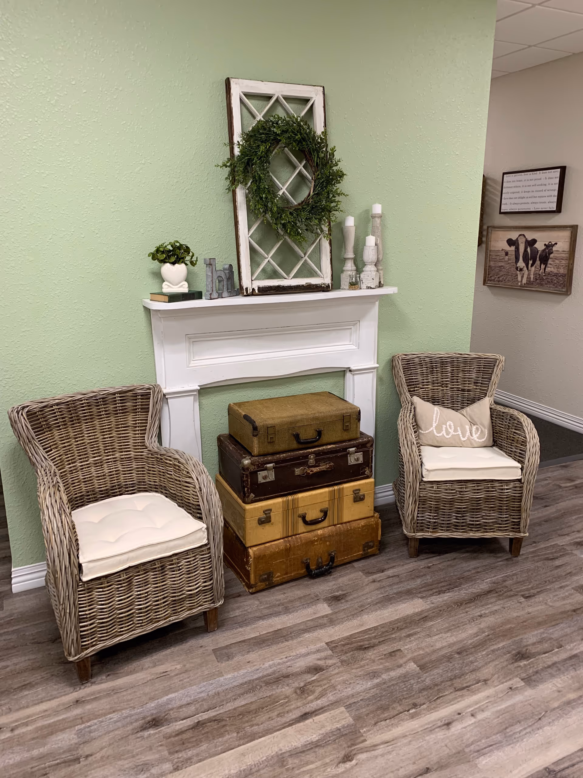 A cozy seating area with two wicker chairs with white cushions, a stack of vintage suitcases between them, and a white decorative mantel on a light green wall. The mantel holds a small plant, decorative letters, a rustic window frame with a green wreath, and three white candlesticks. The floor is wood-patterned, and there are framed pictures on the adjacent beige wall.