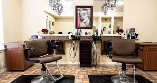 Salon area with two brown styling chairs facing a mirrored counter topped with hair tools and a framed portrait on the wall.