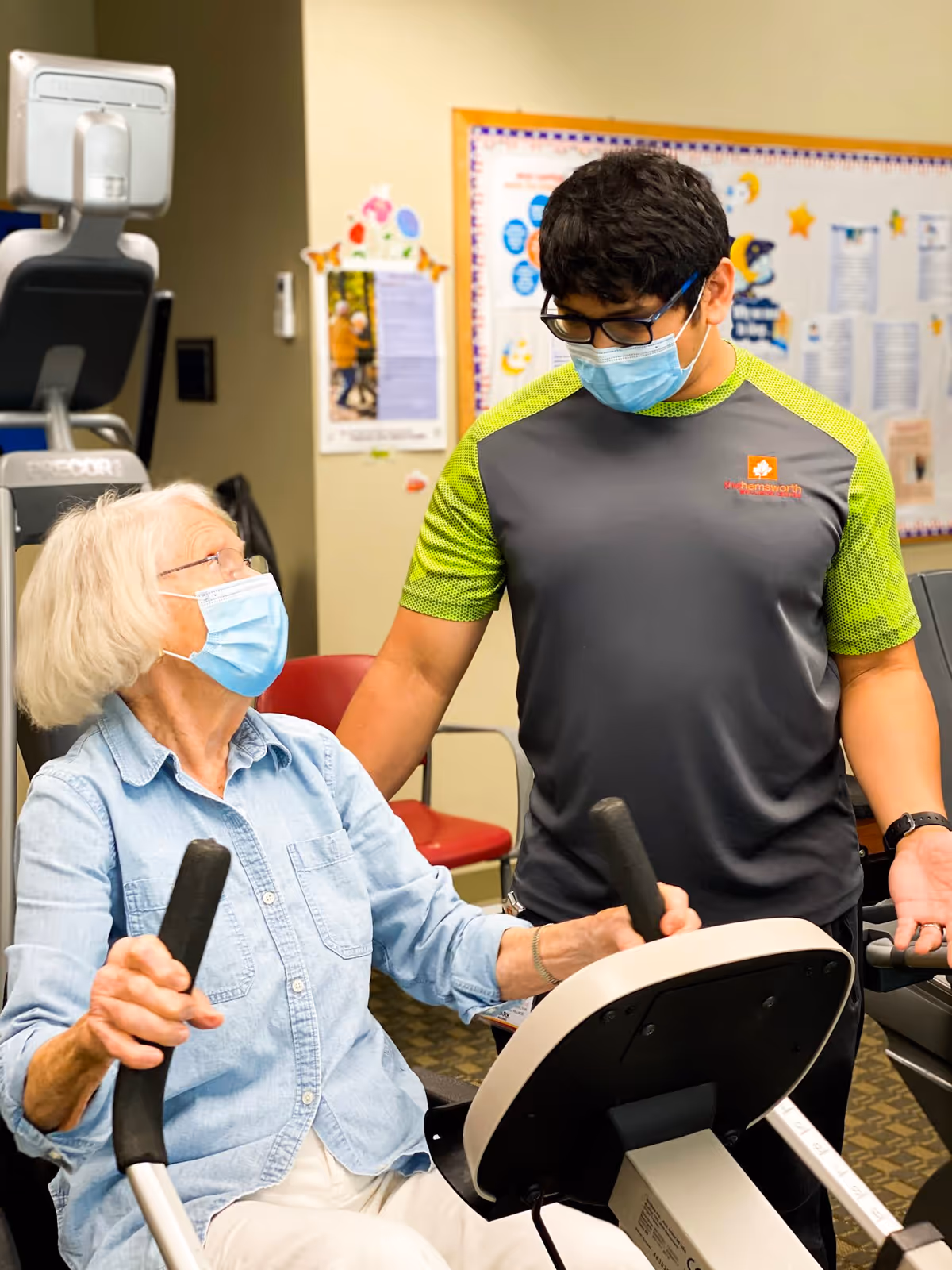 An elderly woman wearing a face mask is using a seated exercise machine while a younger man, also wearing a face mask and a shirt with a logo, stands beside her offering assistance in a fitness room.
