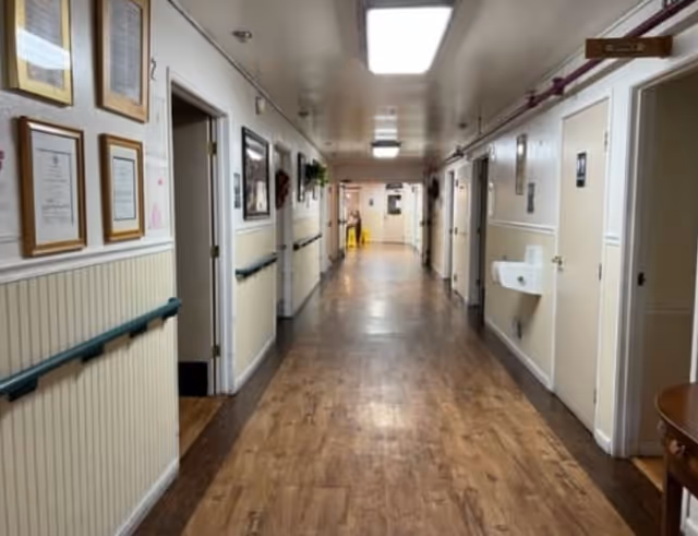 A long, well-lit hallway in a senior living facility with wooden flooring and cream-colored walls. The hallway has multiple doors on both sides, handrails along the walls, framed pictures and certificates hanging on the left wall, and a wall-mounted sink on the right. A caution wet floor sign is visible at the far end of the corridor.