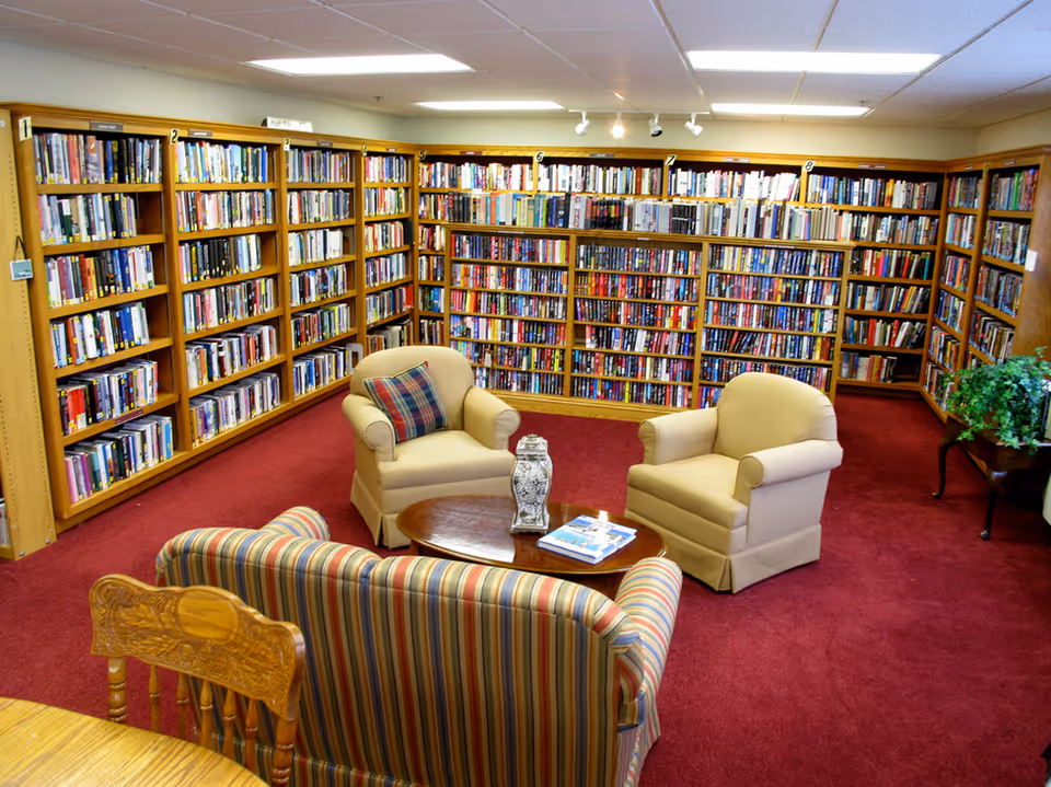 A cozy library room with red carpet, wooden bookshelves filled with books lining the walls, two beige armchairs with a plaid cushion on one, a striped sofa, and a wooden coffee table with a decorative item and a book on it. There is also a wooden chair and a small table with a green plant in the corner.