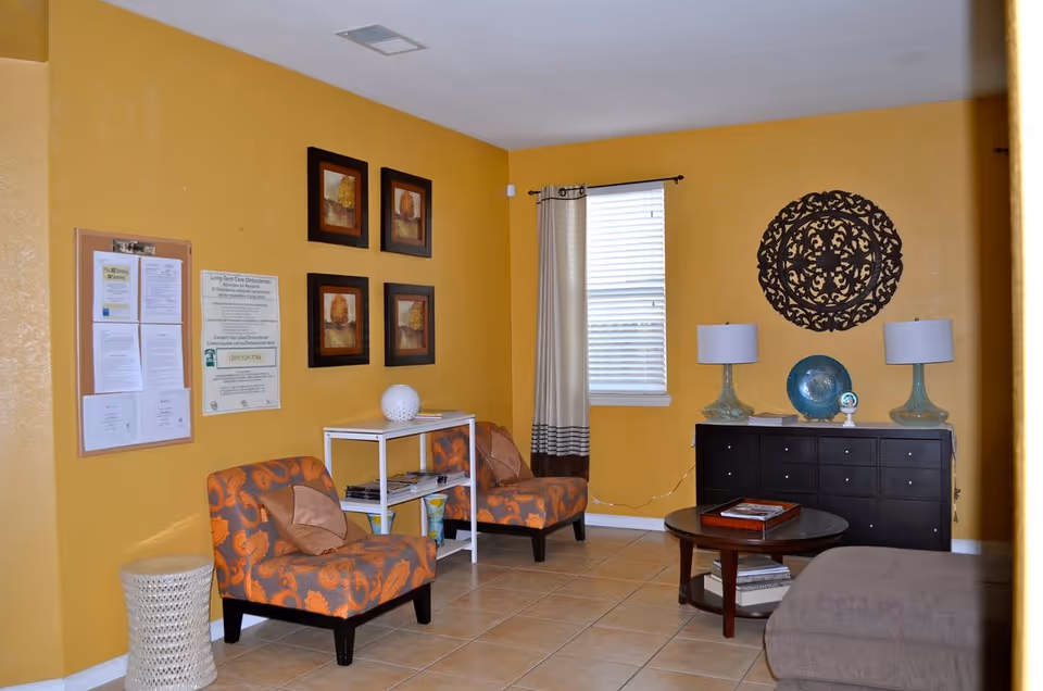 A cozy living room with two orange patterned chairs, a coffee table, a black sideboard with lamps, and mustard-yellow walls.