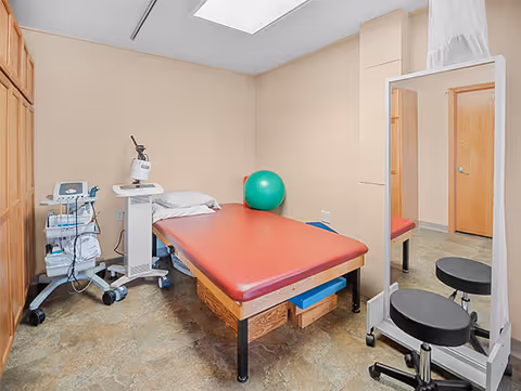A therapy or treatment room with a red padded treatment table, a green exercise ball, medical equipment on a cart, a large mirror on wheels, and a black stool. The room has beige walls, a tiled floor, and wooden cabinets along one wall.