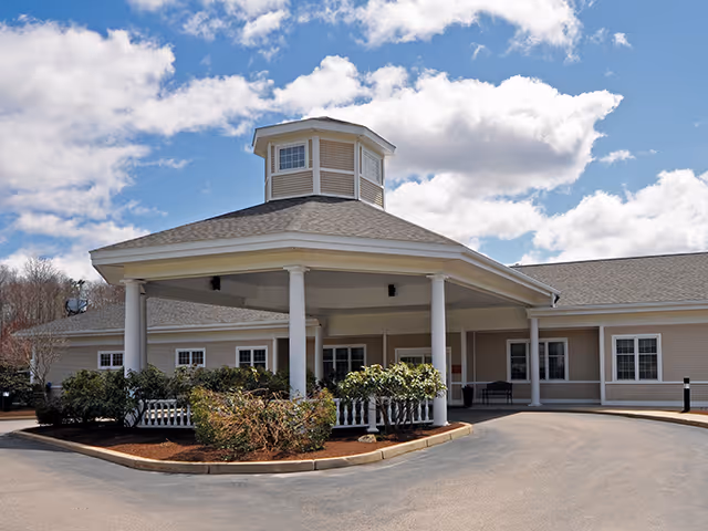 Exterior view of Nemasket Healthcare Center showing the main entrance with a covered drop-off area supported by white columns, surrounded by landscaped bushes and a clear blue sky with some clouds.